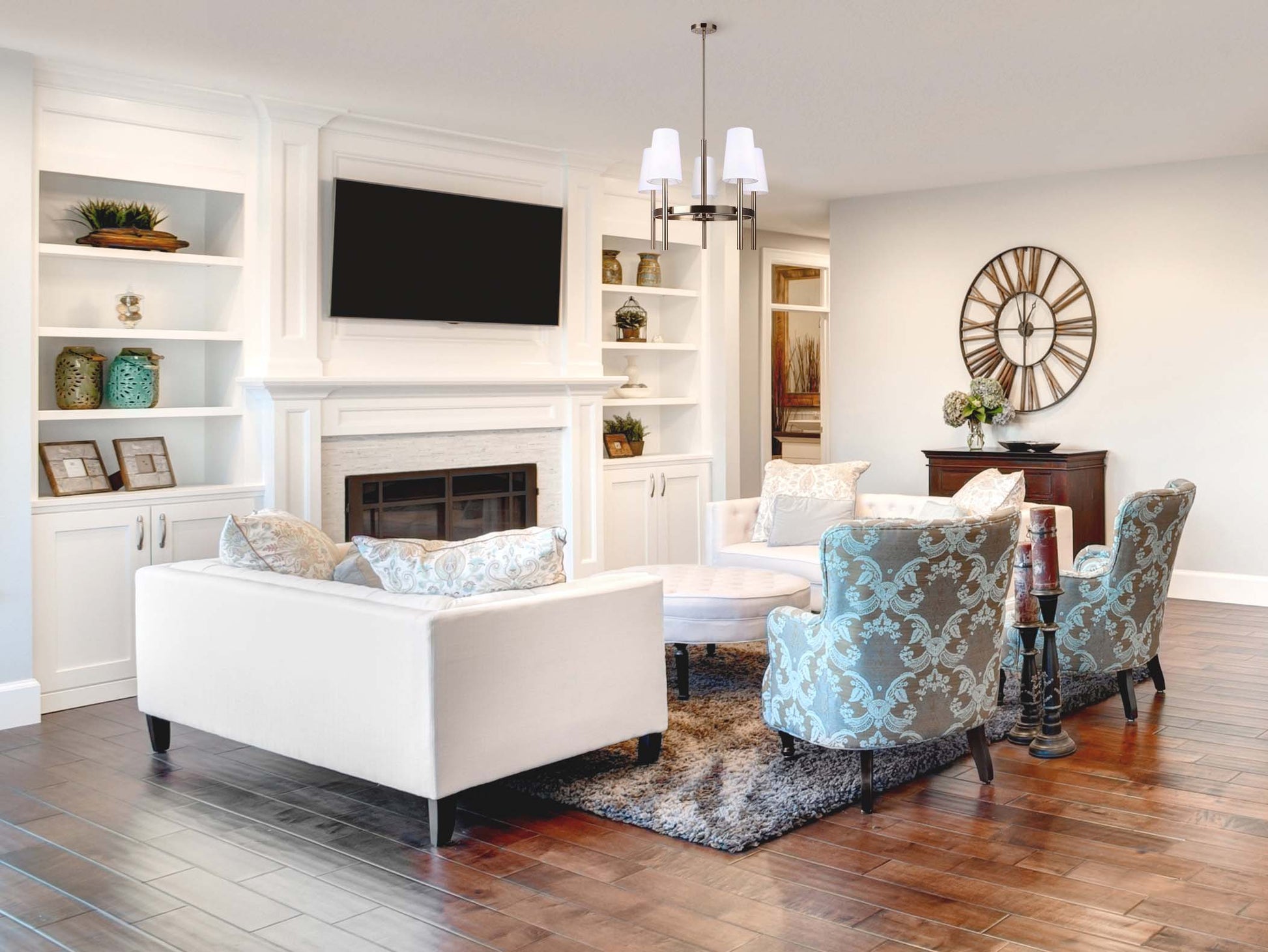 photo of a living room with white walls, tv, gold clock on the wall, two tourquise arm chairs, 5 light chandelier hanging in the middle of the seating area