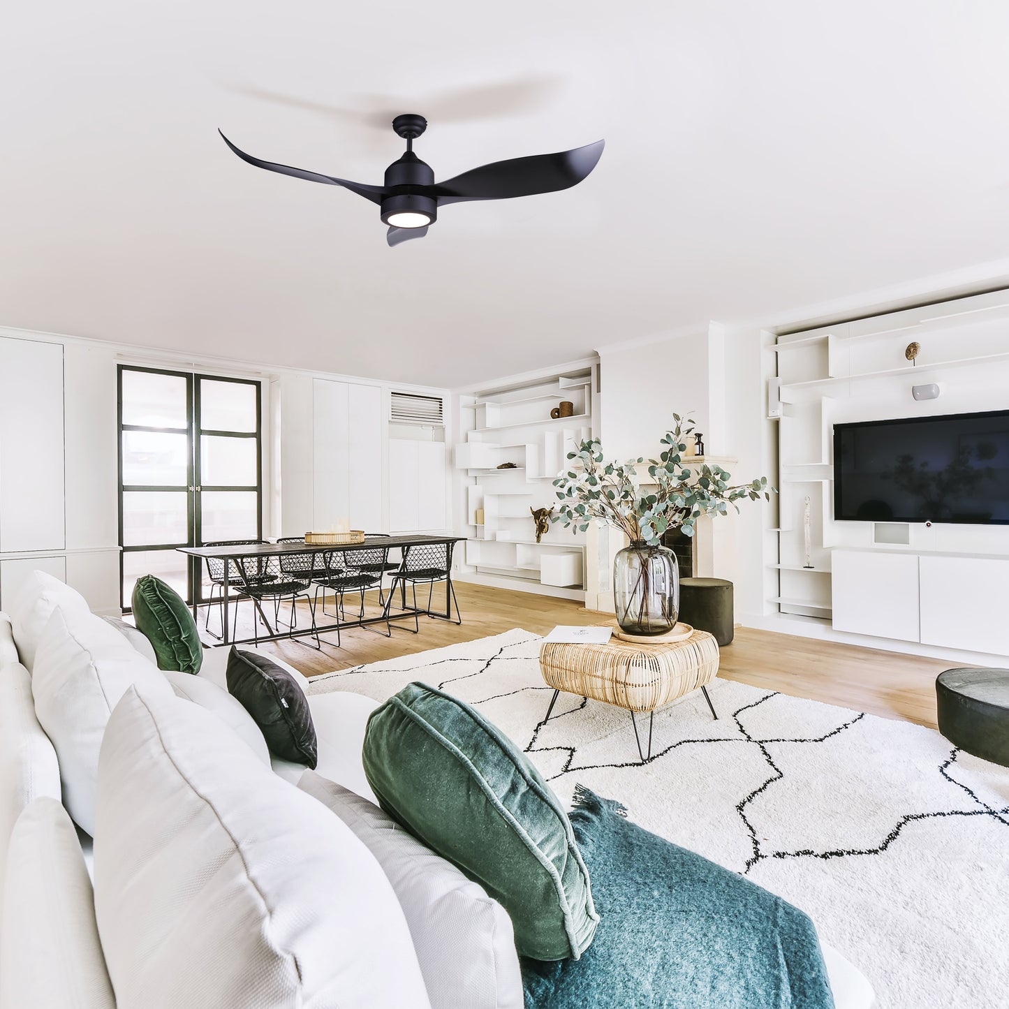 photo of a living room, with white walls, white rug, white couch, black dinning room table in the background. medium size plant in the middle of the seating area, large flat screen tv and a photo of a black 52 inche 3 blade ceiling fan, with a light in the middle. Light is covered with an acrylic lens hanging from the ceiling in the middle of the room